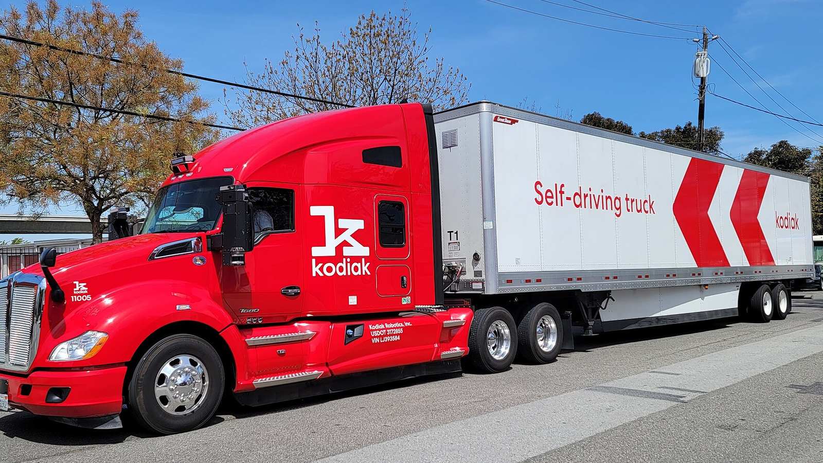A red Kodiak self-driving tractor pulling a white trailer on a road.