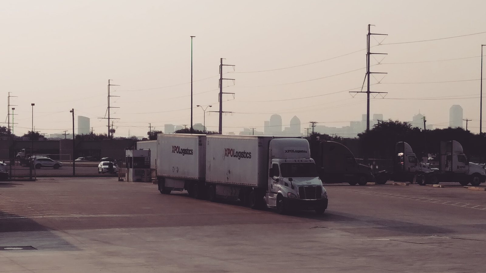 XPO tractors with double trailers in a freight yard, with a city skyline in the distance.