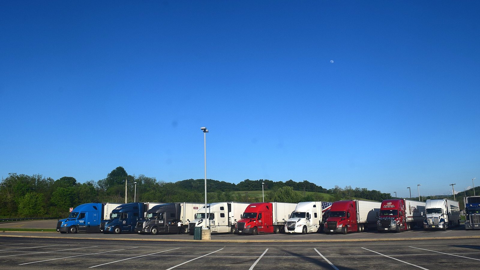 A row of semi-truck tractors parked in a lot beside a highway in the United States.